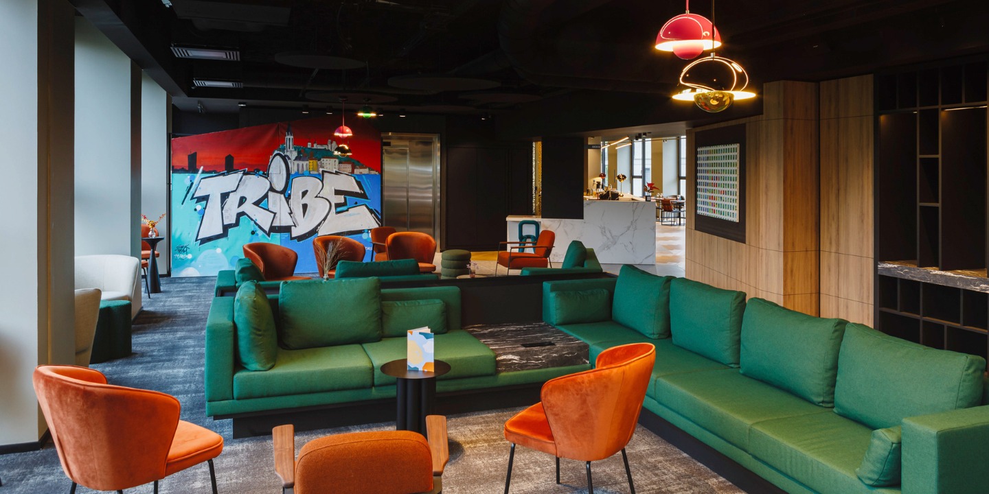 Modern lounge area with green sofas, orange chairs, and a vibrant "TRIBE" graffiti wall, accented by stylish hanging lights.