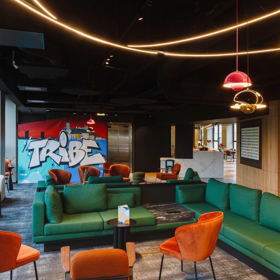 Modern lounge area with green sofas, orange chairs, and a vibrant "TRIBE" graffiti wall, accented by stylish hanging lights.