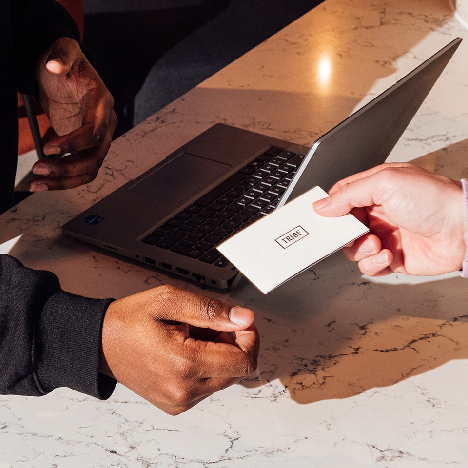A hand passes a white business card with "TRIBE" printed on it to another hand, with a laptop in the background on a marble table.