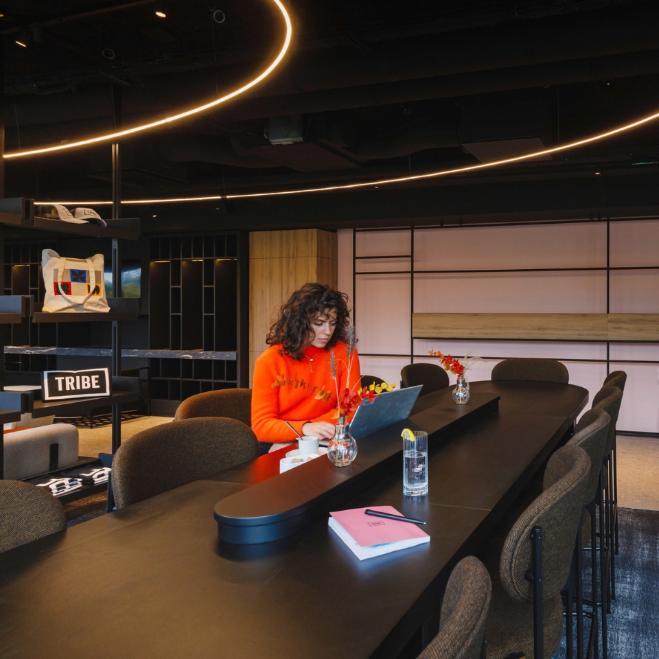 A modern workspace features a long black table with a laptop, plants, and stationery, surrounded by stylish chairs and ambient lighting.