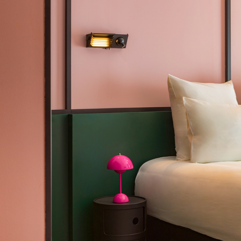 A stylish bedroom corner featuring a pink wall, green accents, plush pillows, and a vibrant pink lamp on a black nightstand.