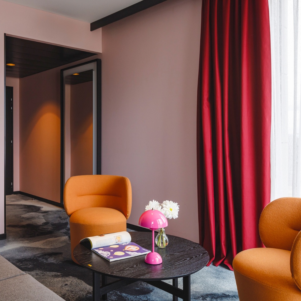 Modern lounge with two orange chairs, a black table, pink lamp, flowers, and red curtains against a soft pink wall.
