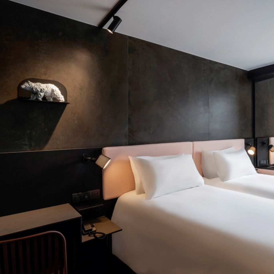A guest room with a twin bed with a pink headboard in front of a black wall, on the left is a small figurine of a bear
