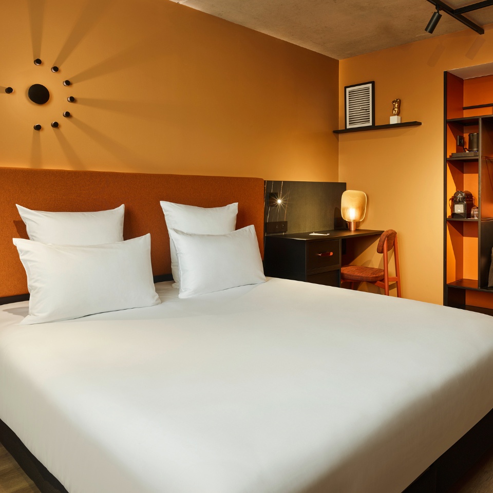 A double bed with an orange headboard and a small black desk next to it, there is a black and orange wardrobe