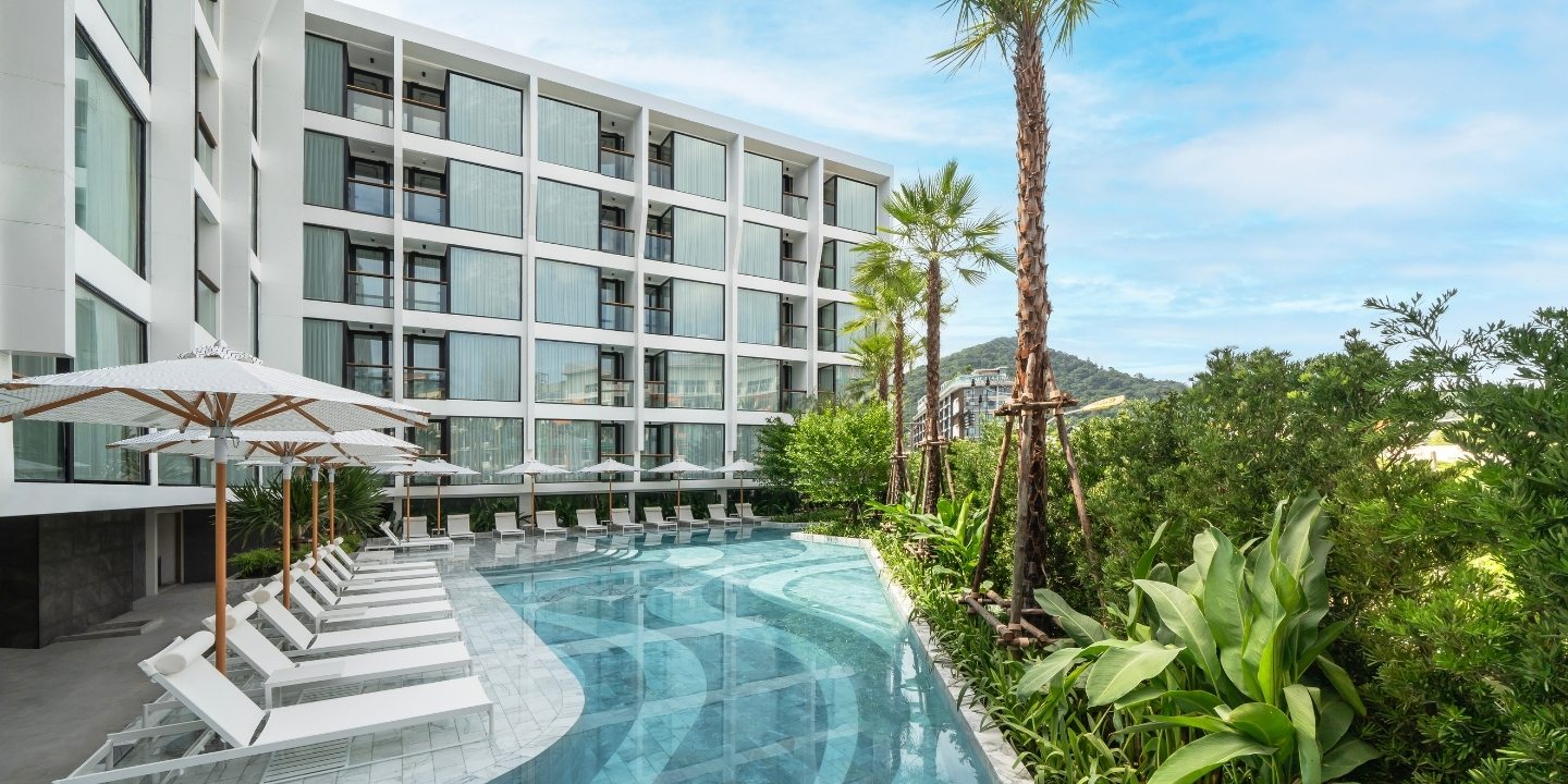 Modern hotel exterior with white, geometric facade overlooking a curvy pool lined with sun loungers and umbrellas. Palm trees and lush greenery add a tropical feel.