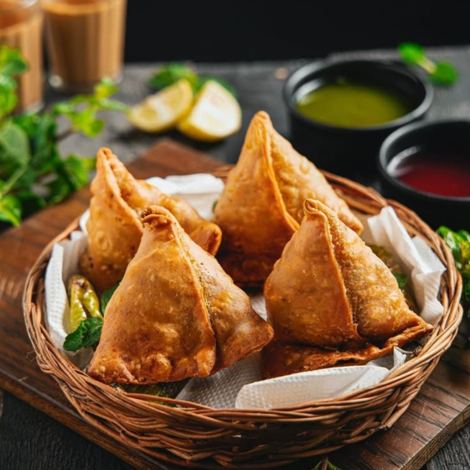 A basket of golden-brown samosas sits on a wooden surface, accompanied by green and red chutneys, lemon wedges, and tea.