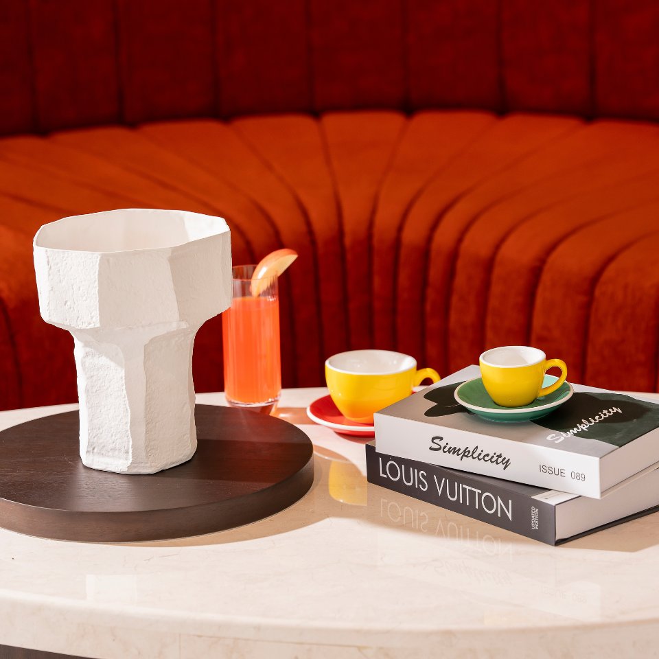 A stylish coffee table with a white textured vase, yellow cups on saucers, a glass of pink drink, and fashion books against a curved orange couch backdrop.