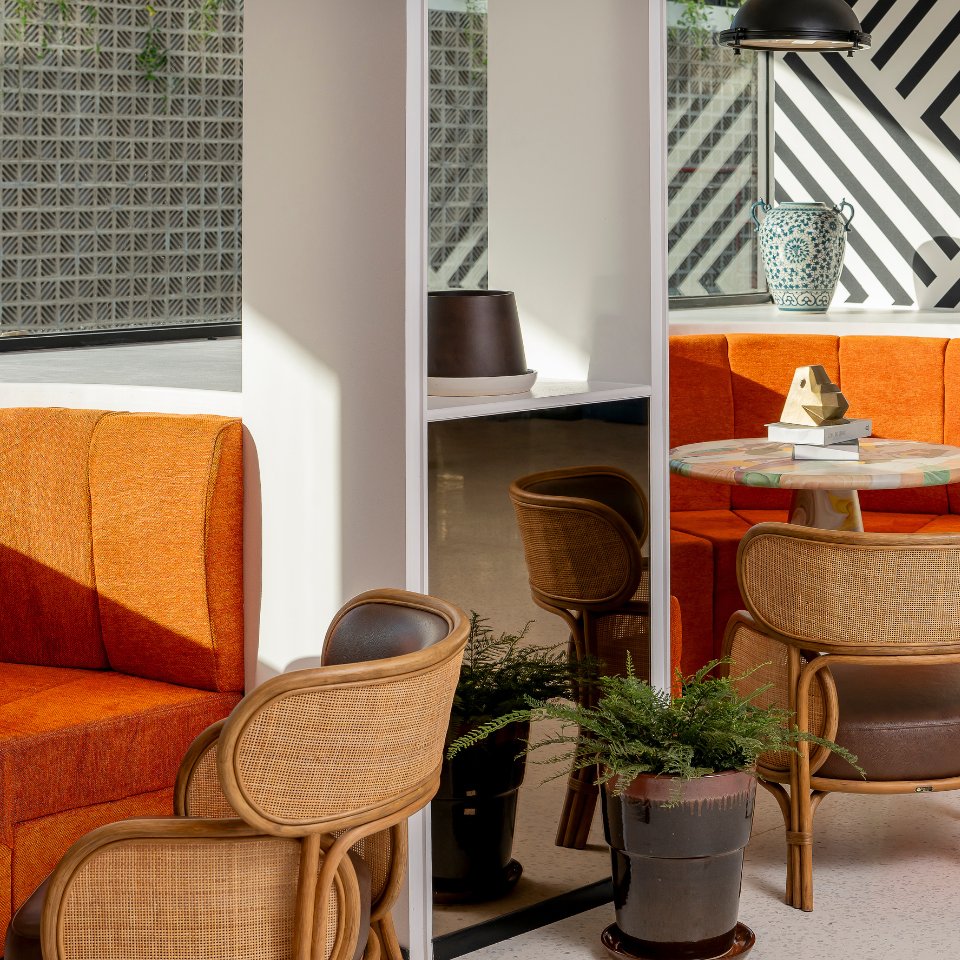 Cozy seating area with orange booths and wicker chairs around a colorful table. A plant, lamp, and ceramic vase add decor. Sunlight and patterns create a warm, modern vibe.