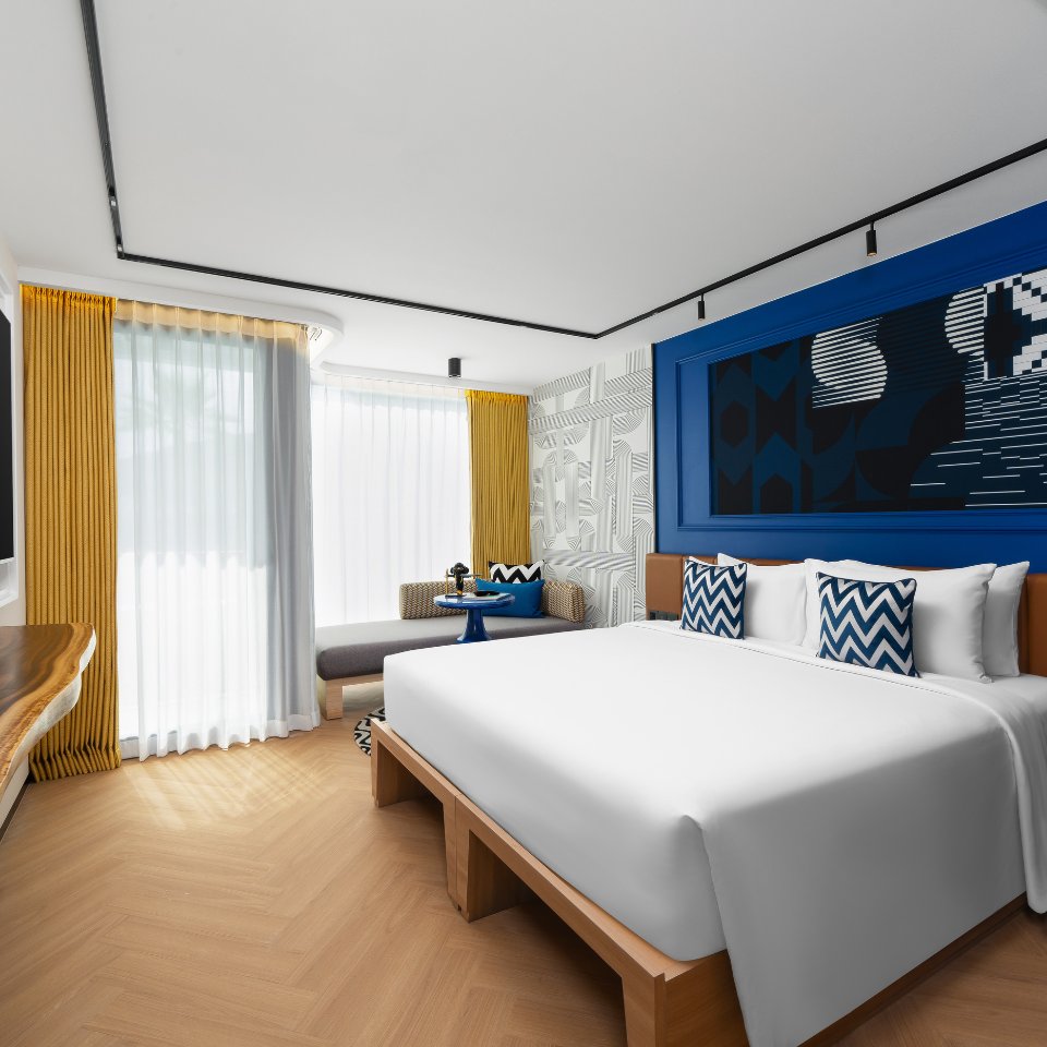 Modern hotel room with a large white bed, blue and white zigzag pillows, vibrant blue artwork, yellow curtains, and a cozy window seat. Bright, inviting atmosphere.