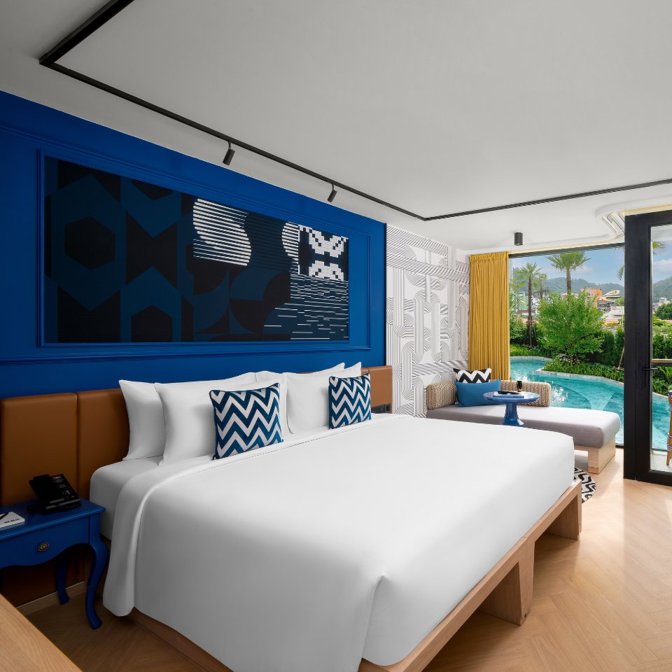 Modern hotel room featuring a king bed, blue accents, patterned cushions, and a cozy seating area overlooking a pool and garden.