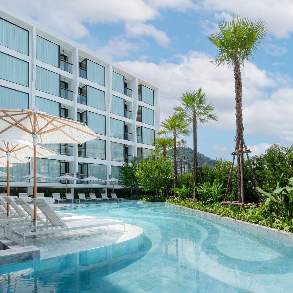 Modern hotel exterior with a curving, clear blue swimming pool. White lounge chairs and parasols line the pool, set against palm trees and green foliage.