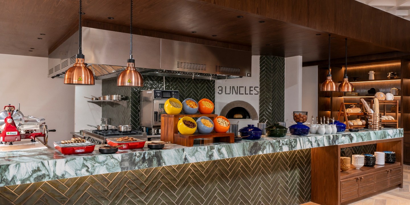 A modern kitchen with a green herringbone tile counter, colorful bowls of nuts, copper lamps, and a wood-fired oven. The mood is warm and inviting.
